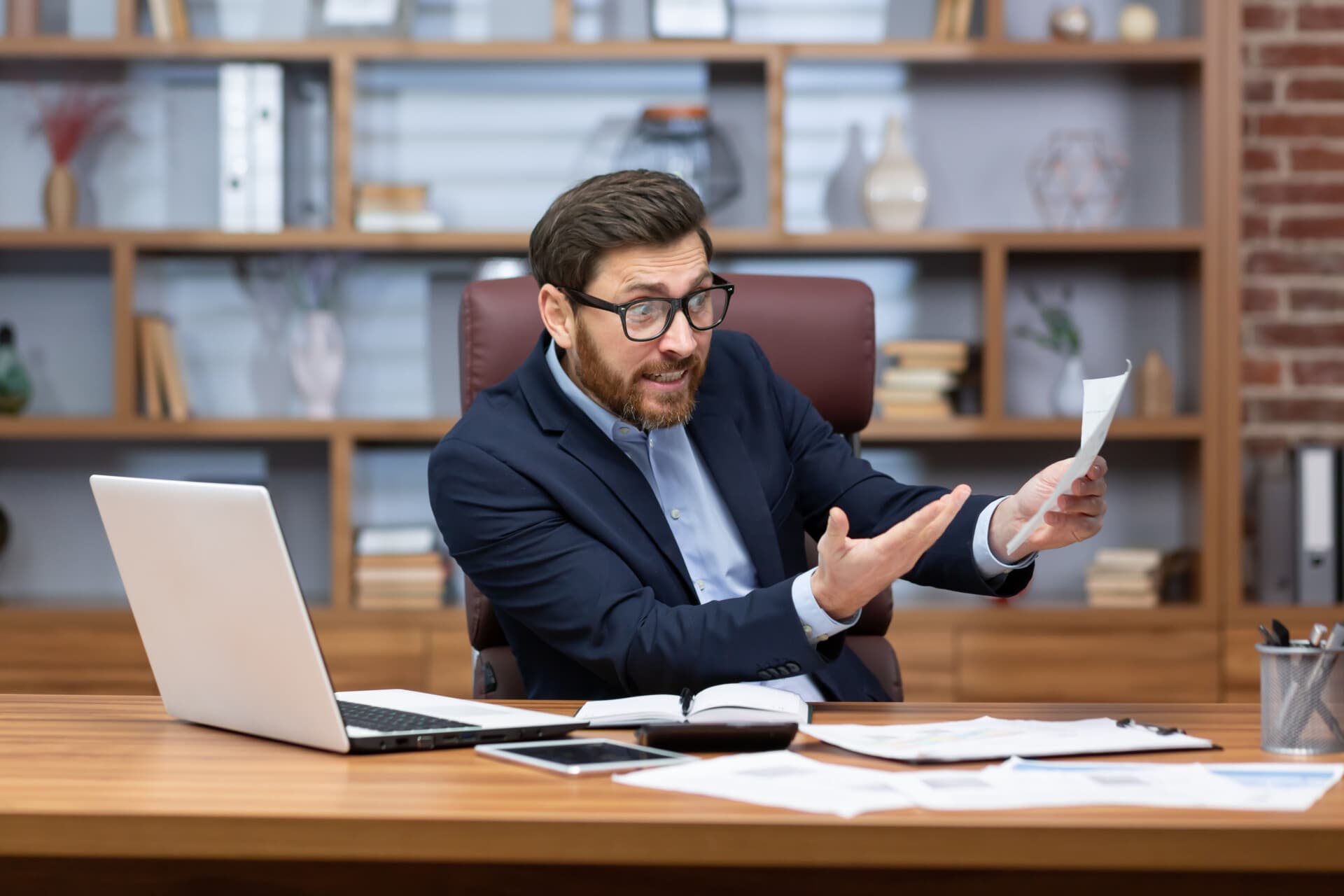 Stock photo of a stressed accountant at his desk, gesturing in frustration while reviewing a paper amid scattered documents and a laptop